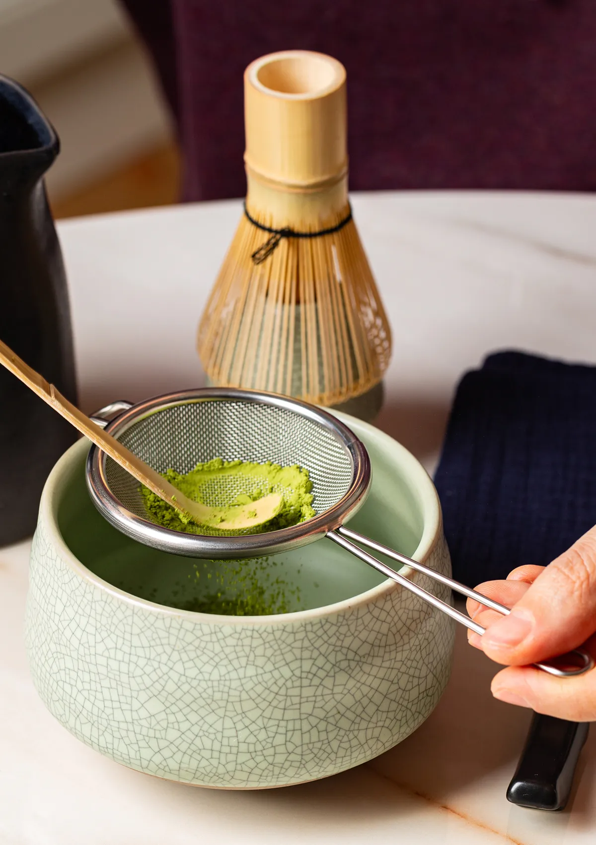 A tea bowl and tea urn filled with matcha tea placed on a bamboo saucer. Matcha and sweets float in the tea bowl, while the calm green scenery and warm light of an andon (paper lantern) can be seen in the background of the tea room.