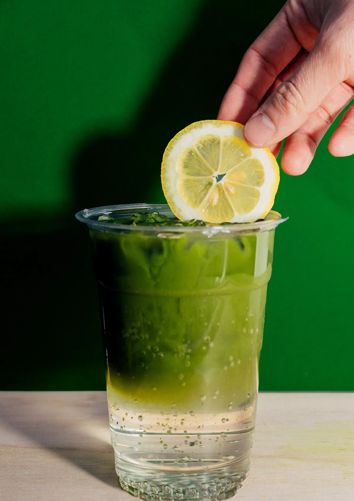 Matcha drink poured into a clear glass. A hand adding a lemon slice to the rim of the glass. Refreshing matcha soda with ice and carbonation.