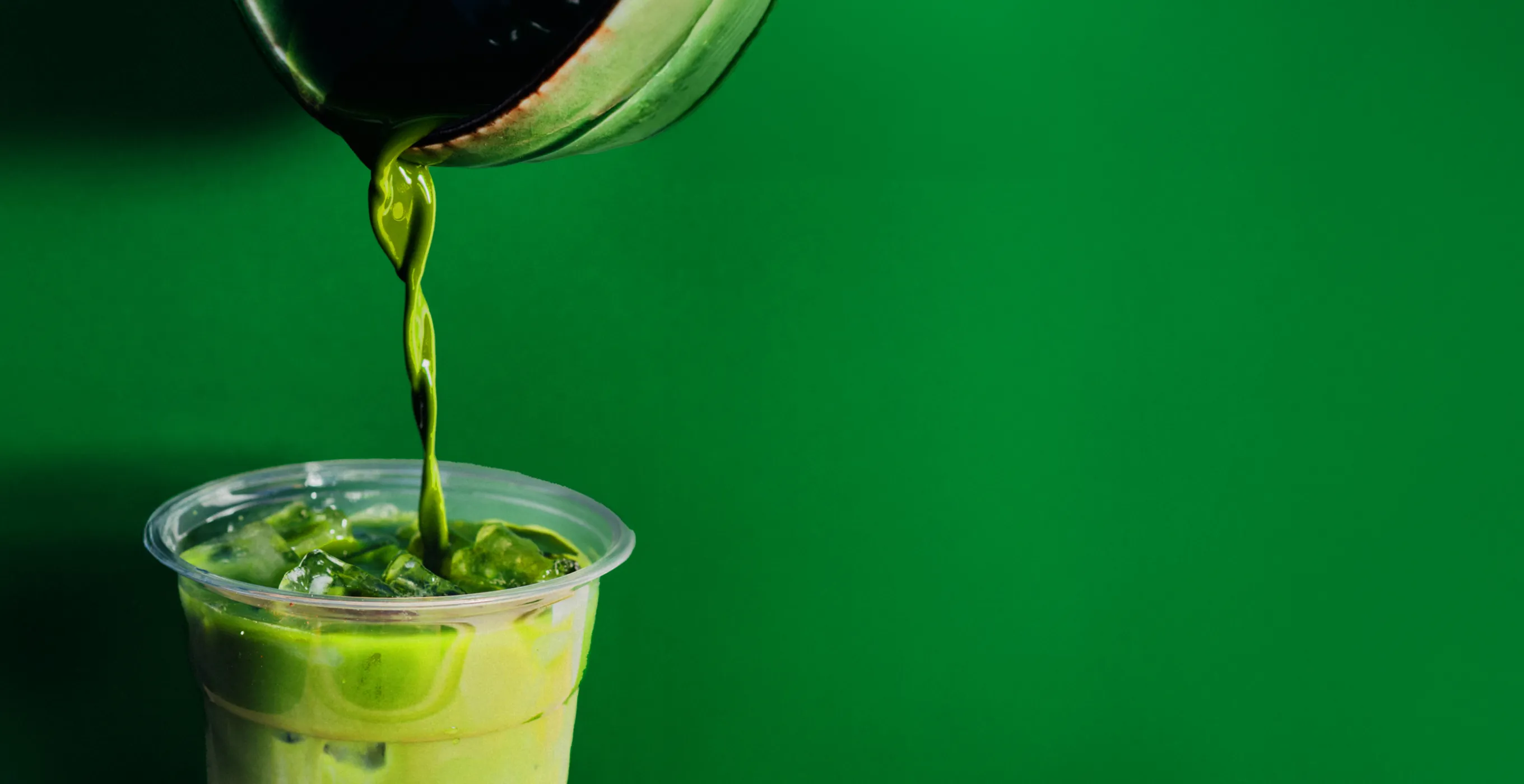 A bright matcha drink poured from a tea cup into a clear cup. This stylish matcha drink photo captures the moment the green liquid flows as it is poured into a cup filled with ice.