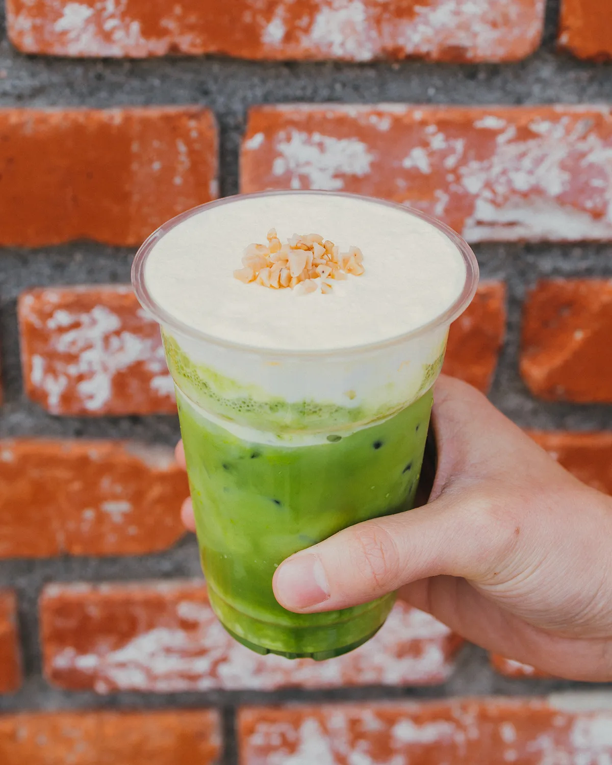 A photograph of a matcha latte held in the hand against a brick wall. The bright green matcha drink in a transparent cup is topped with fluffy white milk foam and creamy nuts.