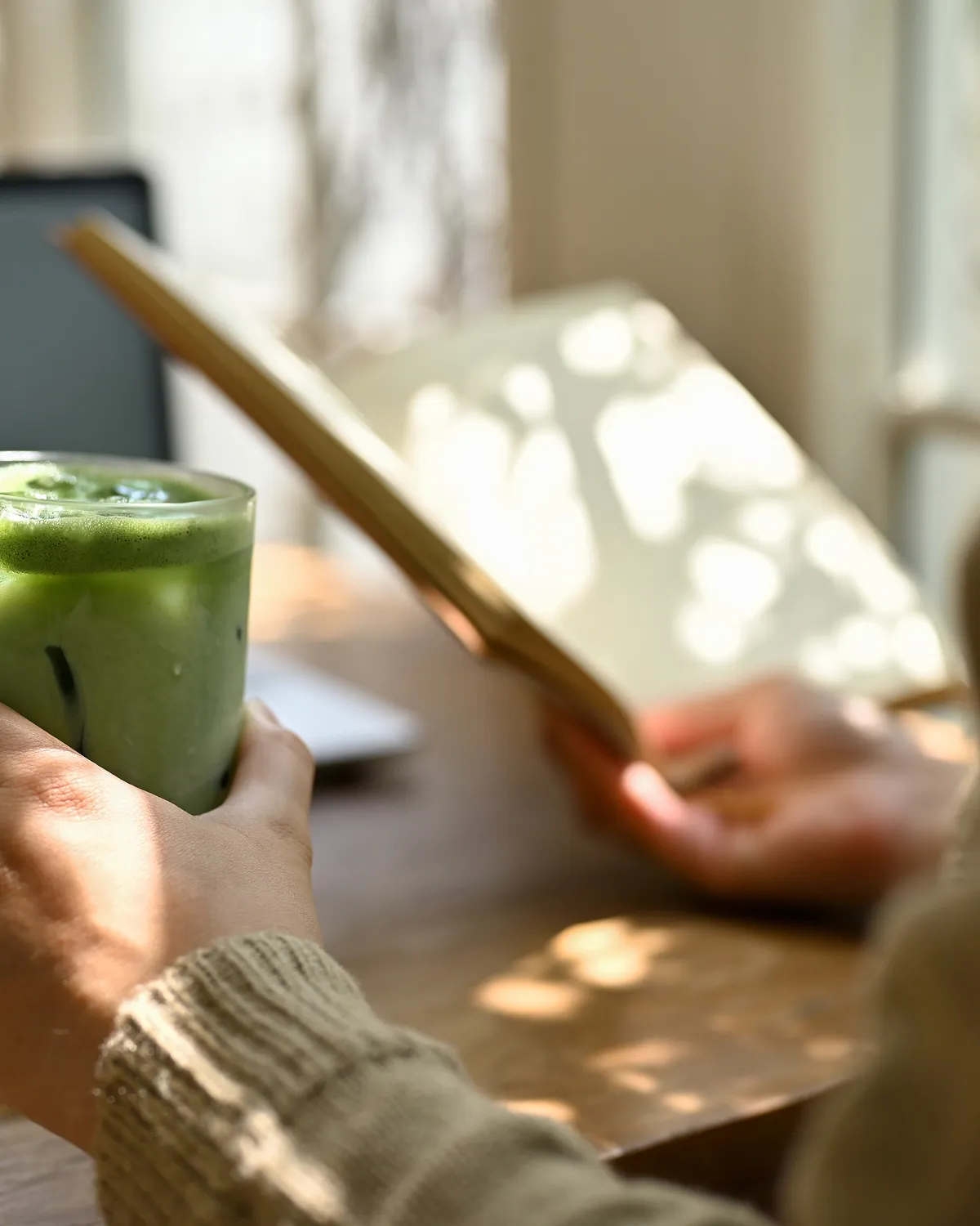 A person in a gray sweater holds a green matcha drink in a glass while reading a tablet or book in the other hand. Soft sunlight shines on the wooden tables, creating a relaxing atmosphere.