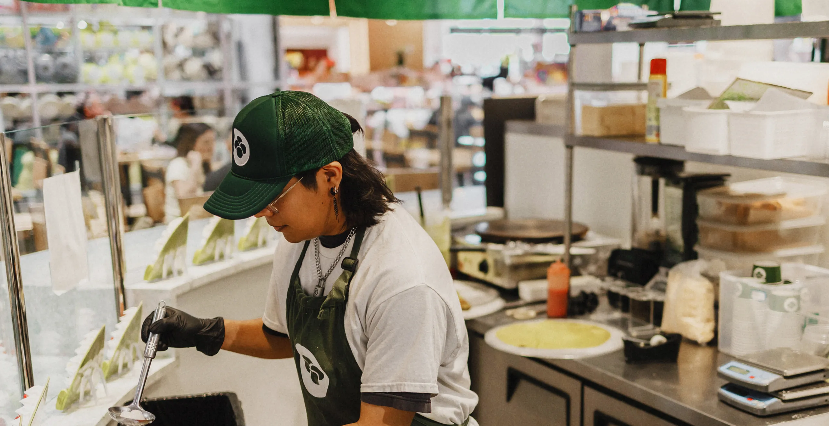 A staff member wearing a green hat working in the store's kitchen. Wearing a white T-shirt and black apron, they appear to be working over a customer service counter. A view of a matcha specialty store or café.